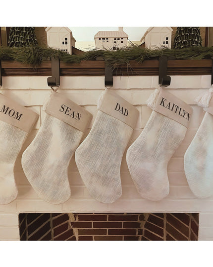 Rustic wooden mantel with black metal stocking holders displaying cream burlap Christmas stockings personalized with names...