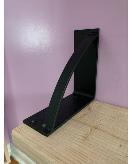 Black metal triangle shelf bracket "The Brie Bookshelf Bracket" by Industrial Farm Co mounted on natural wood against purp...