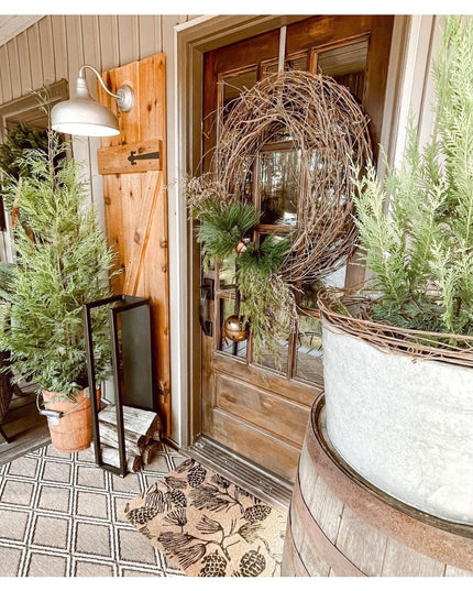 Black metal Toby Firewood Holder by Industrial Farm Co displayed on rustic porch with evergreen branches and wicker wreath...