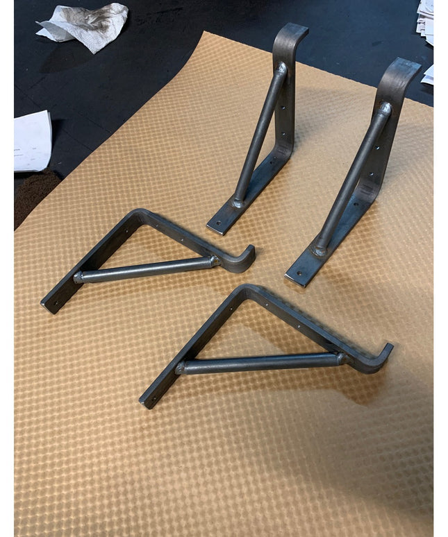 Four black metal Norwich Support Brackets by Industrial Farm Co displayed on textured surface, showing curved wrought iron...