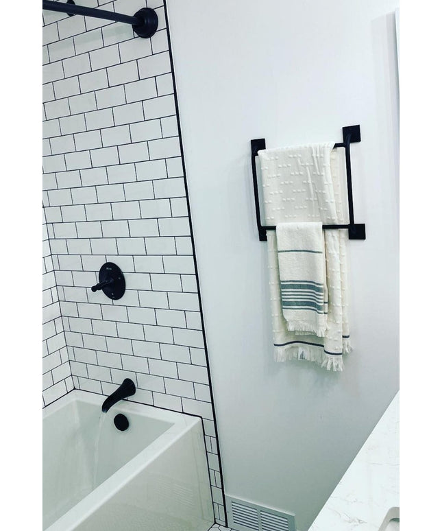 Black metal Parker Hand Towel Holder ladder mounted on white bathroom wall with cream towels, next to white subway tile sh...