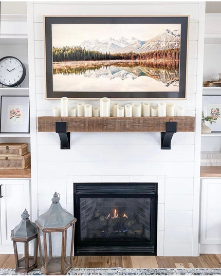 Kudlick Farmhouse black metal mantel brackets supporting rustic wood beam shelf with white candles above white brick firep...