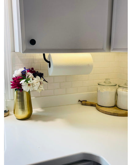 Black metal Hyde Hall Paper Towel Holder mounted under white kitchen cabinet with white paper towel roll on marble countertop