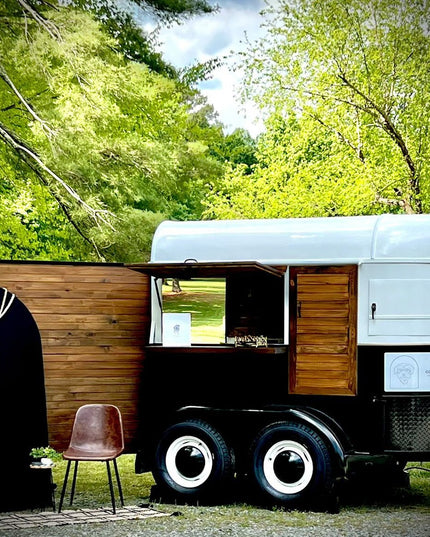 Vintage white and black mobile coffee trailer with wood paneling parked among green trees, featuring service window and ou...