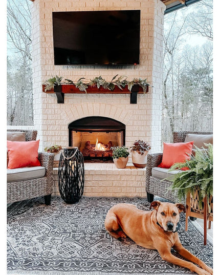 Black Kudlick Farmhouse Mantel Brackets supporting wooden mantel shelf with plants above brick fireplace in outdoor living...