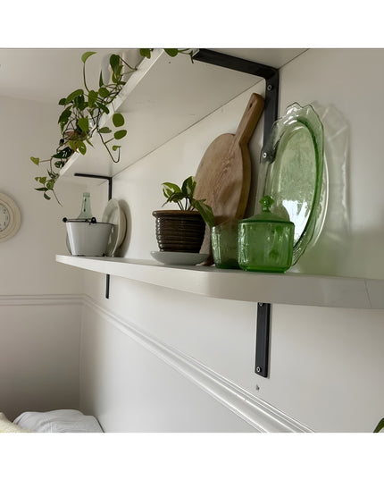 Black steel L-shaped shelf brackets supporting white floating shelf with green glassware, plants, and wooden cutting board...
