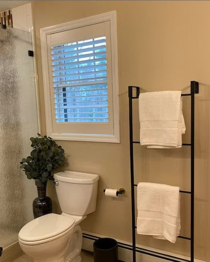 The Veronica Modern Blanket Ladder by Industrial Farm Co in black metal frame holding white towels in bathroom with toilet...