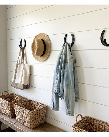 Black metal horseshoe wall hooks mounted on white shiplap wall holding hat, bags, and denim jacket with wicker storage bas...