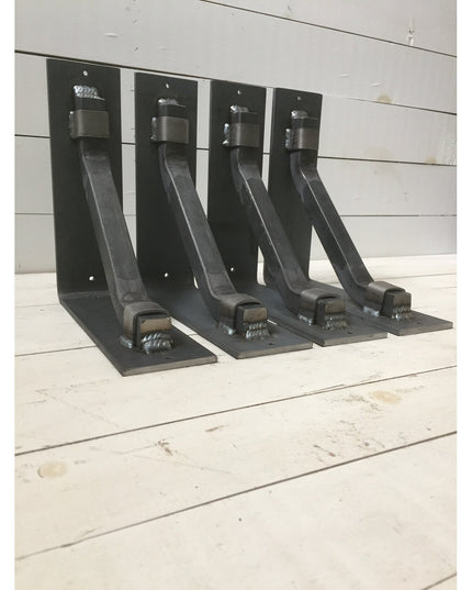Four black metal Hornell Industrial Shelf Support brackets from Industrial Farm Co displayed on white wooden planks, showi...