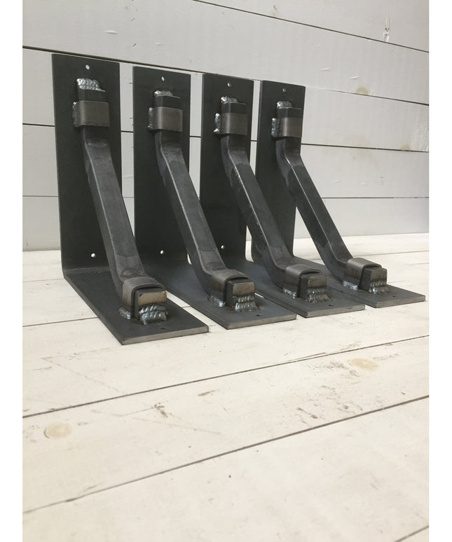 Four black metal Hornell Industrial Shelf Support brackets from Industrial Farm Co displayed on white wooden planks, showi...