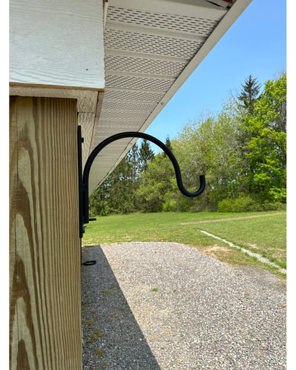 Black wrought iron Gregory Shepherd Hook by Industrial Farm Co mounted on wooden post under building overhang with green f...