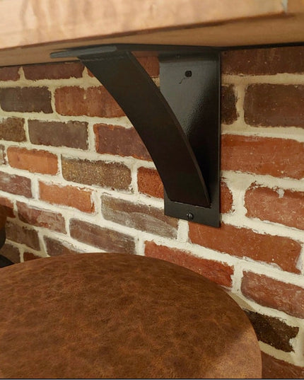 Black steel Albany 4-inch industrial shelf bracket with curved support bar mounted under wooden shelf against exposed bric...