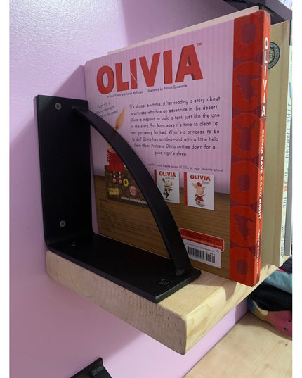 Black triangular metal bookshelf bracket from Industrial Farm Co holding an Olivia children's book on wooden shelf against...