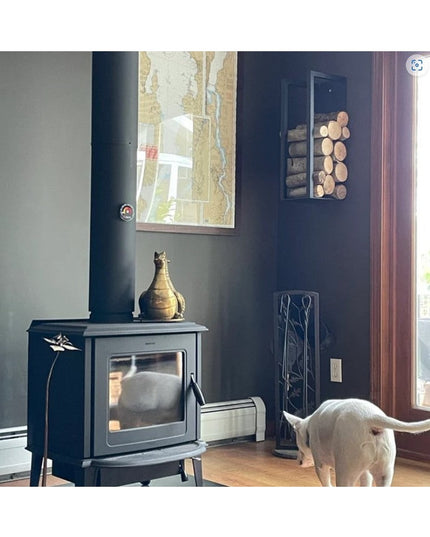 Black metal Toby Firewood Holder by Industrial Farm Co mounted on wall next to wood burning stove, holding stacked firewoo...