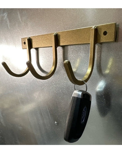 Rustic brass-colored metal key rack with 3 curved hooks mounted on stainless steel wall, holding black key fob from Indust...