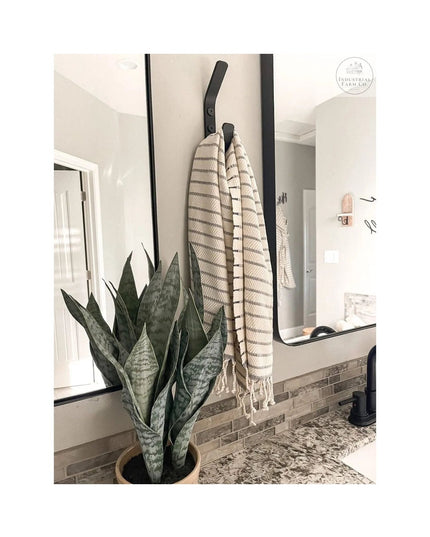 Black metal Henrietta 1" Wide Double Hook mounted on bathroom wall holding striped towels, with snake plant and granite co...