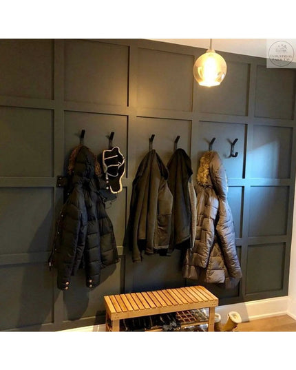 Black metal double hooks mounted on dark gray paneled mudroom wall holding winter coats and jackets with wooden bench belo...