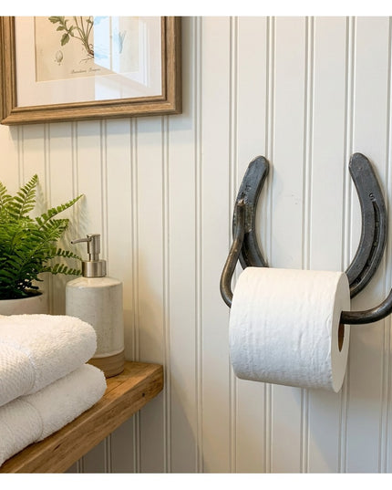 Rustic black horseshoe toilet paper holder mounted on white beadboard wall in farmhouse bathroom with towels and plant dec...