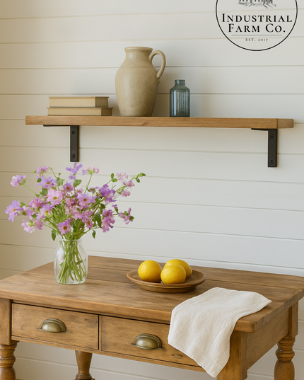 Wooden table with a vase of flowers, lemons, and a towel, with a shelf above featuring a pitcher and bottles against a white paneled wall.