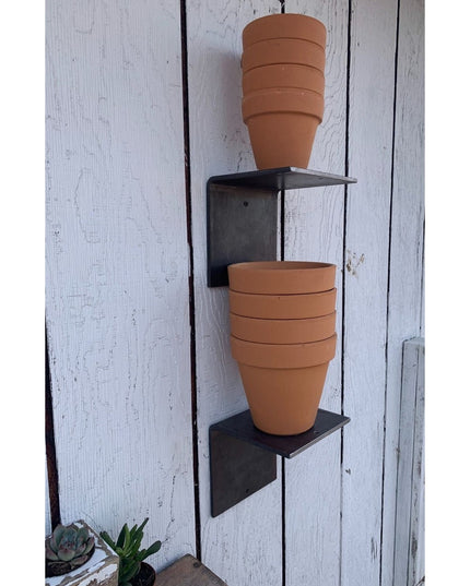 Heavy duty black steel L shelf brackets mounted on white wood fence holding stacked terracotta clay pots - Industrial Farm...