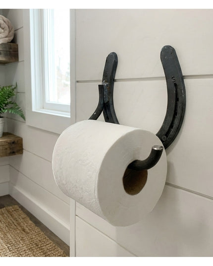 Rustic black horseshoe toilet paper holder mounted on white shiplap bathroom wall with farmhouse decor and floating wood s...